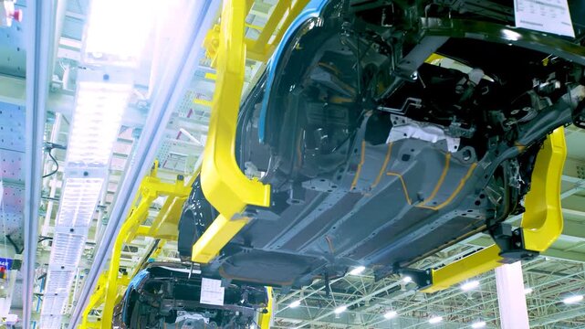 Automotive factory assembly line shows multiple unfinished vehicle car chassis being built with robotic arms, body hanging on overhead yellow conveyor system structure advanced manufacturing equipment