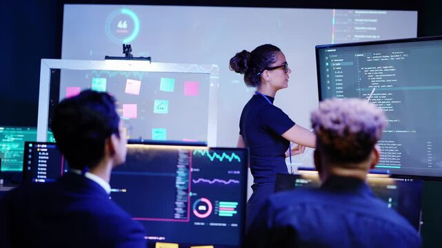 Latina woman writes on a tablet then reaches out to point at Python code on a large monitor as an Asian colleague gestures and a Black teammate holding a pen watches