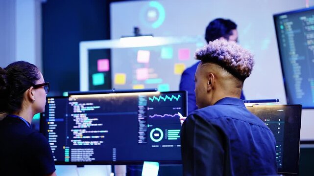 Latina engineer and Black developer seated at code monitors watch an Asian businessman present at a sticky note board and wall screen, she smiles as he turns back to the room