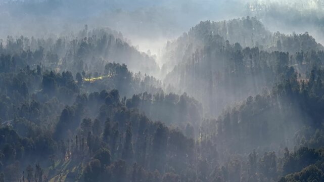 Aerial view of misty layered hills with sunbeams streaming through forested slopes at Mount Bromo.