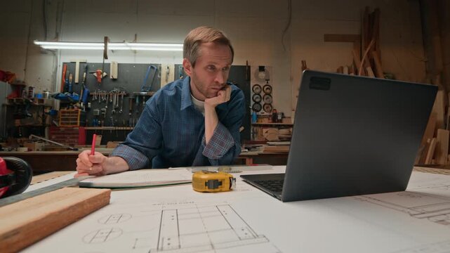 Middle aged Caucasian carpenter drafting blueprints and watching laptop screen in woodworking workshop. Perfect for craftsmanship education and furniture design marketing