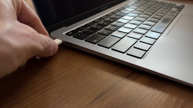 Hand plugging white USB C cable into silver laptop side port to charge, wide low angle view on wooden desk with copy space