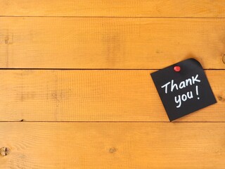 Black adhesive note with handwritten thank you text pinned on a yellow wooden wall. Flat lay composition with copy space, ideal for gratitude, business appreciation, and greeting card design © Eka