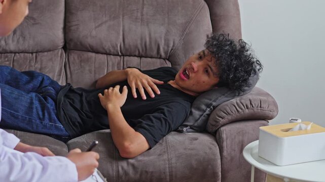 Doctor taking notes while young man on couch holding chest in somatic distress during PTSD therapy
