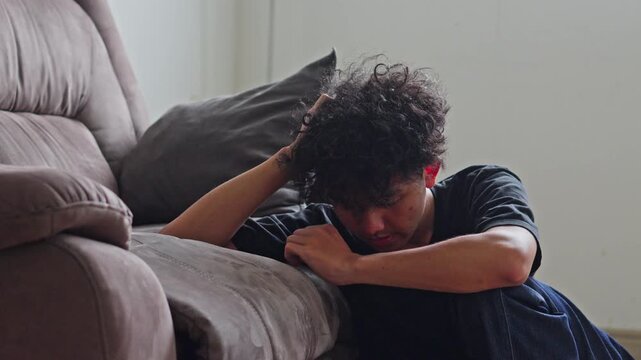 Young man sits slumped on floor next to couch hiding face in profound distress during PTSD episode