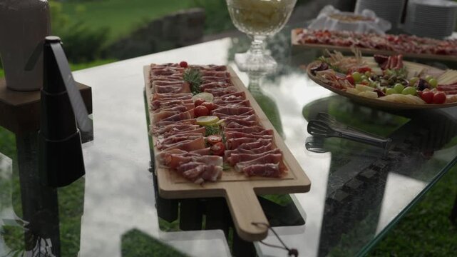 Long wooden board of thinly sliced cured ham garnished with cherry tomatoes, next to a cheese platter on a glass buffet table on a lawn at a wedding reception.
