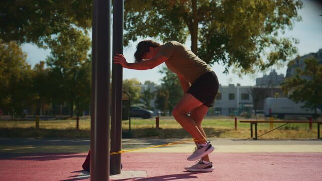 Man performs controlled resistance band leg kickbacks against a workout pole during outdoor fitness training.