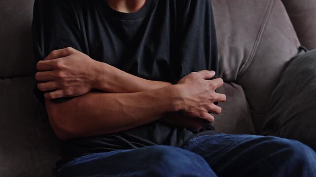 Young man sitting on couch tightly hugging himself with crossed arms portraying severe PTSD chills