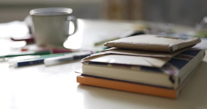 Cinematic close-up of a stack of books and kraft paper envelopes on a light table, colorful markers and a ceramic mug in soft focus. Cozy creative workspace, shallow depth of field, natural light.