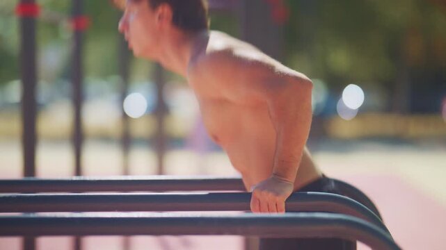 Shirtless man performs tricep dips on parallel bars during a slow motion outdoor calisthenics workout in a sunny fitness park.