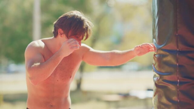 Man throws jab and cross punches at a heavy bag during outdoor boxing training in a sunny park, shown in slow motion.