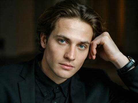 Close up portrait of a handsome young man with blue eyes and brown hair wearing a black suit jacket and shirt with a black leather watch strap