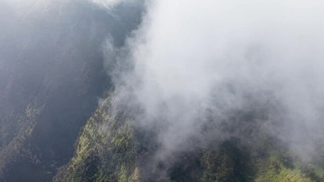 Mist curls around jagged mountain peaks. Green slopes fade into hazy depth below at Wonokerto