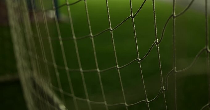 Cinematic close-up of a football goal net with detailed mesh texture against a blurred green soccer field. Shallow depth of field creates an atmospheric sports background with soft lighting.