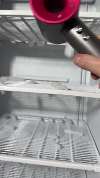 Man defrosts a freezer using a hairdryer's hot air, ice chunks falling and melting, puddles on floor, with towels and bucket, candid authentic life domestic maintenance task at home.