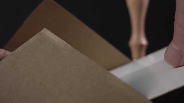 Hands slide a white card into a brown kraft envelope and close the flap. A wooden wax seal stamp with a brass head stands in the background on a dark set.