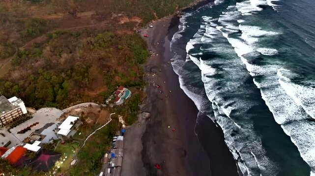 Aerial view of Parangtritis Beach with dark volcanic sand, crashing waves, and coastal resorts in Kecamatan Kretek, Daerah Istimewa Yogya, Indonesia.