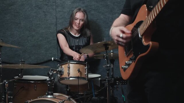 Band jam session with female drummer and electric guitarist playing together, woman following groove and smiling while musicians rehearse instrumental music in studio.