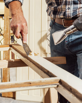 Carpenter using heat gun and wood scraper removes paint from old wooden window. Construction industry, carpentry. Restoration. 
