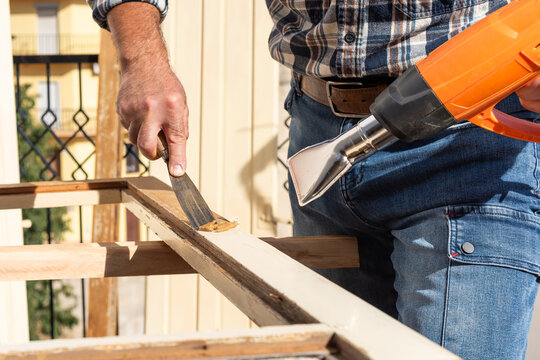 Carpenter using heat gun and wood scraper removes paint from old wooden window. Construction industry, carpentry. Restoration. 