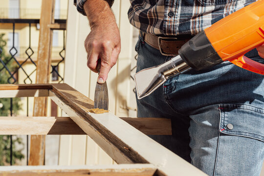 Carpenter using heat gun and wood scraper removes paint from old wooden window. Construction industry, carpentry. Restoration. 