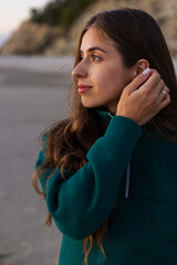 Woman in teal jacket tucks hair behind her ear beside a rocky coast at golden hour © Тарас Белецкий