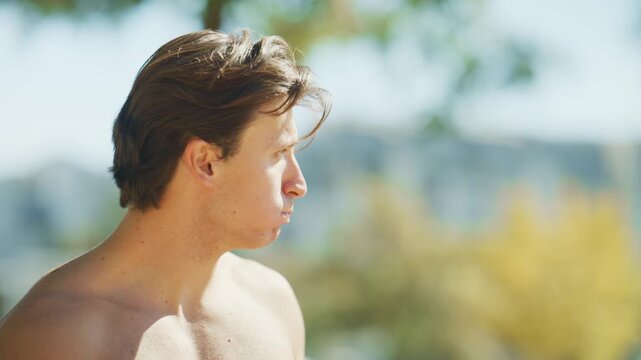Shirtless man drinks water from a sports bottle after an outdoor fitness workout, hydrating and recovering in summer sunlight.