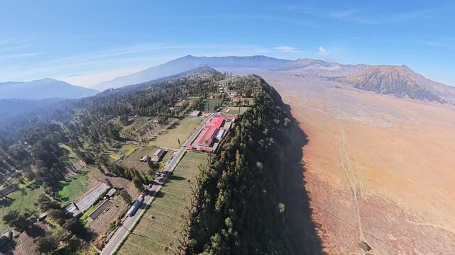 Drone fisheye aerial view of village on steep crater rim and vast volcanic plain under clear sky.