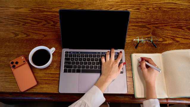 Female hands taking notes beside laptop with blank screen on wooden desk, overhead view. Online courses, business planning and productivity tools concept.