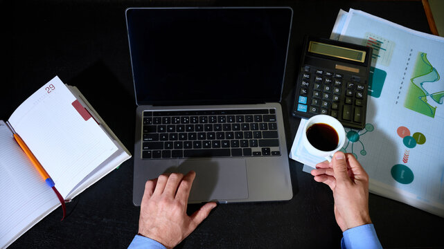 Male hands using laptop and holding espresso beside calculator and printed charts, overhead view. Accounting services, financial planning and business analytics concept.