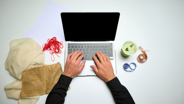 Clothing designer typing on laptop beside fabric samples, thread and measuring tapes, overhead view. Fashion entrepreneurship, textile sourcing and tailoring courses concept.
