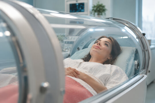 A woman lies on a bed inside a clear hyperbaric chamber, with her hands resting over a pink blanket. Medical equipment and a bright clinical room are visible in the background