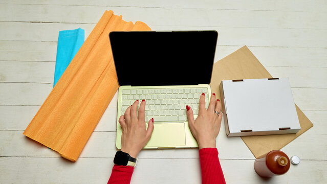 Woman using laptop beside packaging box and colorful wrapping paper on white wooden table, overhead view. E-commerce, order fulfillment and handmade business concept.