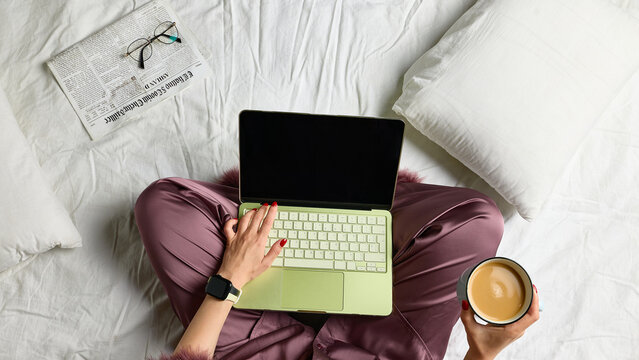 Pajama-clad woman using laptop and holding coffee while sitting cross-legged on bed, overhead view. Flexible schedules, digital subscriptions and home leisure concept.