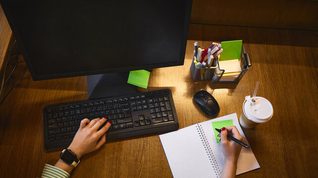 Office worker typing and writing notes beside desktop monitor with blank screen, overhead view. Administrative services, task management and employee training concept.