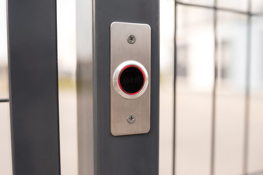 Close up of modern metal gate intercom button with red ring element on grey steel fence post outdoors, selective focus daytime shots, commercial secure property entry background, industrial design