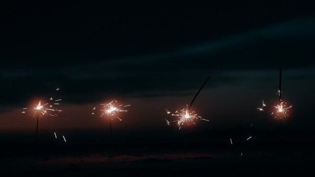 Multiple Sparklers Burning and Sparkling Row in Sand on Beach at Night