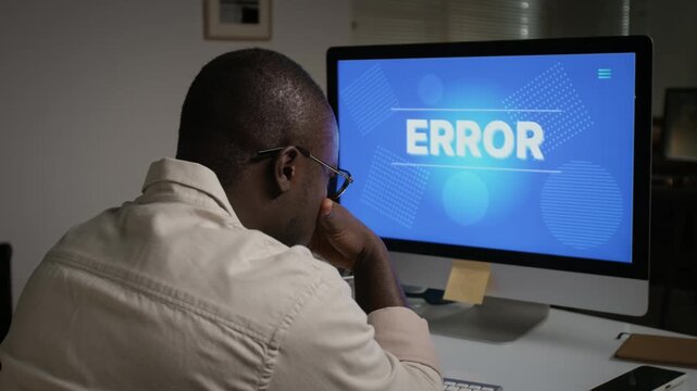 African American young adult man facing computer screen with error message, feeling frustrated by system failure. Suitable for IT support, cyber security, and technical troubleshooting projects