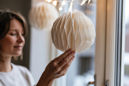 In a charming indoor environment, a young woman delicately holds a honeycomb-style paper lantern, emphasizing creativity and ambiance with a backdrop of soft lighting and joyful expressions.