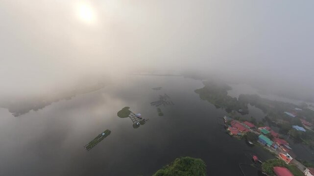 Aerial fisheye view misty dawn blankets calm waters and distant village rooftops. Sunlight pierces fog