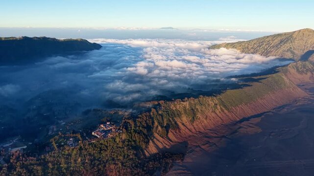 Misty clouds roll over rugged volcanic terrain. Sunlight glows softly on distant hills and slopes at Bromo