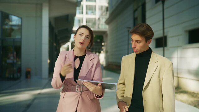Two business professionals walk outside a modern building, discussing information displayed on a digital tablet.