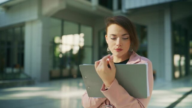A focused businesswoman in a pink blazer carefully examines documents on a clipboard outdoors. She appears concentrated, working in a modern urban environment.