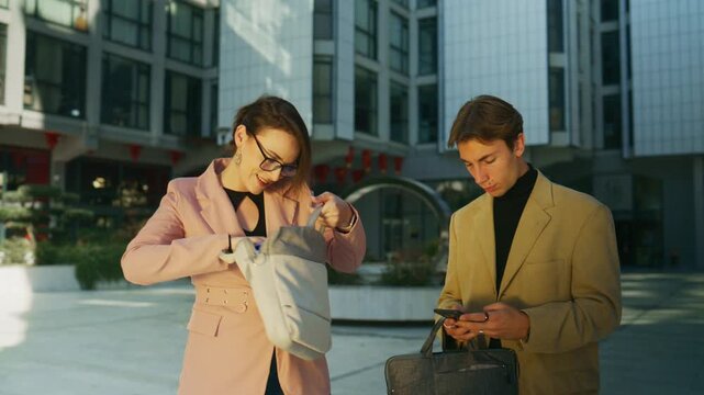 A businesswoman searches her bag while a young man checks his mobile phone in a modern urban environment.
