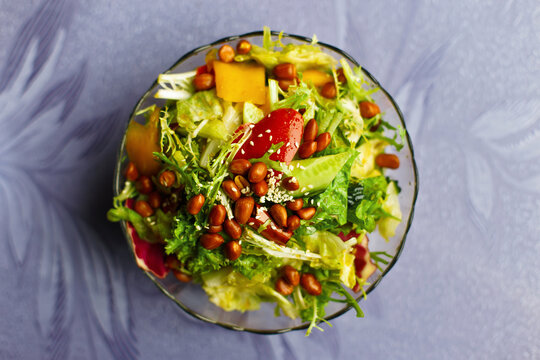 Fresh healthy Asian salad with leafy greens, crunchy peanuts, cucumbers, peppers, and sesame seeds served in a glass bowl on a purple background. Top view close-up of delicious vegetarian food.