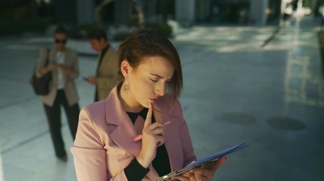 A focused businesswoman diligently writes on a clipboard outdoors, deep in thought. She meticulously reviews documents, concentrating on her planning and strategy in a modern urban setting.