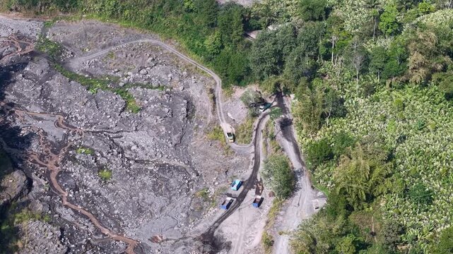High angle drone view of volcanic ash plain, winding road and trucks beside green jungle