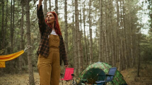 Red-haired woman breaks firewood and stacks kindling beside a tent at a pine forest campsite while preparing for a campfire.