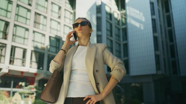 A confident businesswoman in sunglasses talks on her smartphone in a modern urban district. She stands poised and professional, managing business communication outdoors.