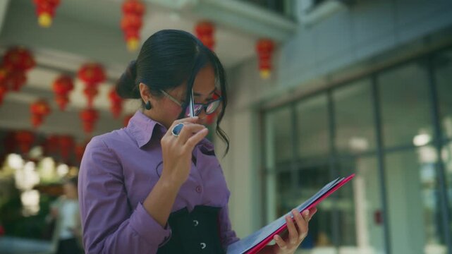 A focused young Asian businesswoman reviews documents outdoors, holding a pen and a red folder. She concentrates on her work in an urban setting adorned with traditional Chinese lanterns.